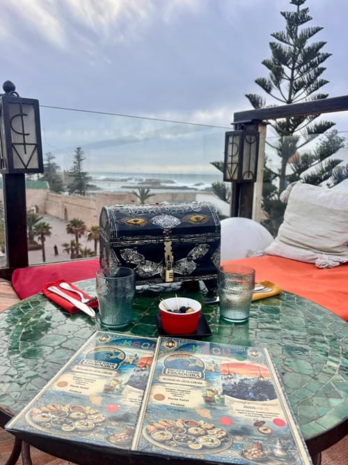 Dîner en bord de mer au Riad Eucalyptus avec vue océan, coffre décoratif, menus et palmiers à El Ghazouane