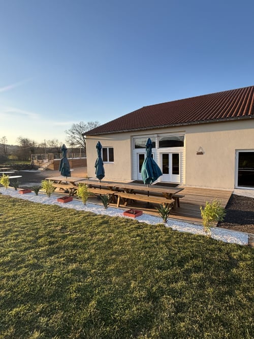 Patio de la salle de réception du Mas de Bannayoux avec tables de pique-nique en bois, parasols bleus et pelouse impeccable à Couteuges, France