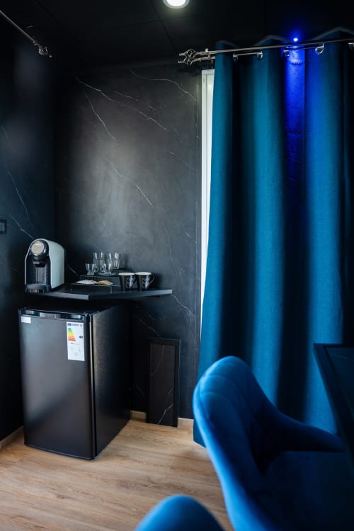 Camera d'hotel moderna con pareti in marmo nero, tende blu, piccolo angolo cottura con macchina da caffè e pavimento in legno presso LHE