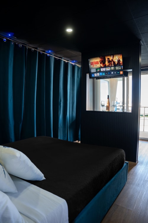Camera moderna con tende turchesi, TV al plasma con Batman, letto nero, cuscini bianchi e vista sul mare - BHE House Boat