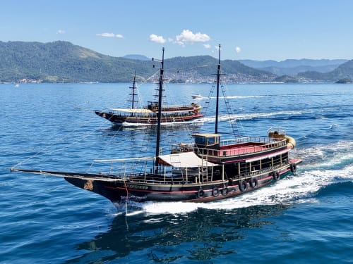 Barcos à vela tradicionais ancorados em águas turquesas com montanhas arborizadas, representando a costa do Rio de Janeiro