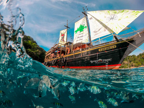 Navio pirata com velas brancas ancorado em águas turquesas com peixes tropicais e selva ao fundo perto do Rio de Janeiro