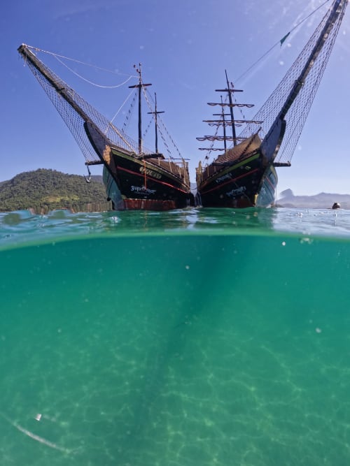 Dois veleiros tradicionais com altos mastros em águas turquesas perto do Rio de Janeiro, montanhas ao fundo