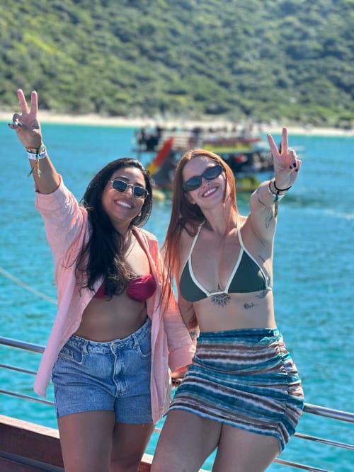 Duas mulheres fazendo sinais de paz em um barco com águas turquesas e costa florestal na Rio Vibes Hostel, Rio de Janeiro