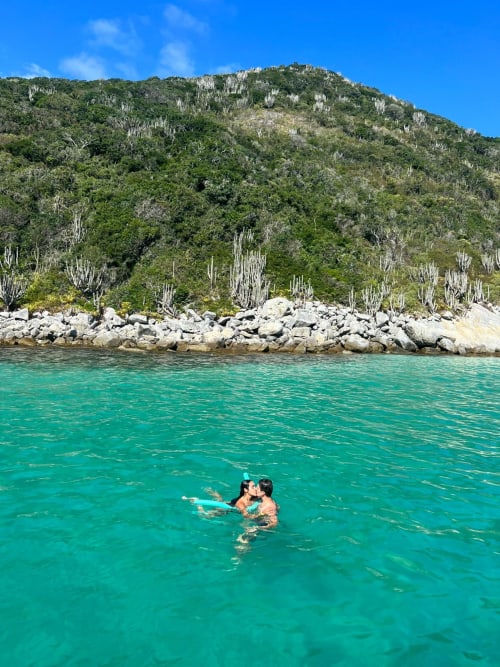 Casal nadando em águas turquesas tropicais perto de uma costa rochosa com vegetação exuberante e cactos.