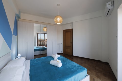 Camera da letto moderna con biancheria blu, armadio specchiato, lampade a sospensione e balcone con vista mare - Appartamento ViOletta Roma