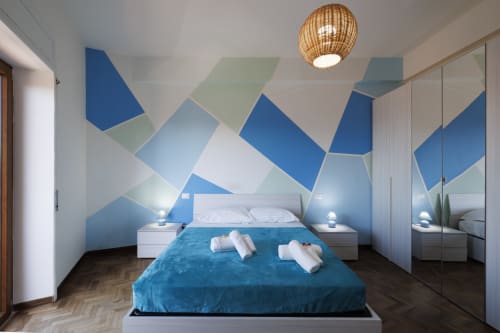 Camera da letto moderna con arte geometrica blu a muro, letto bianco con coperta blu, guardaroba specchiato e plafoniera in vimini