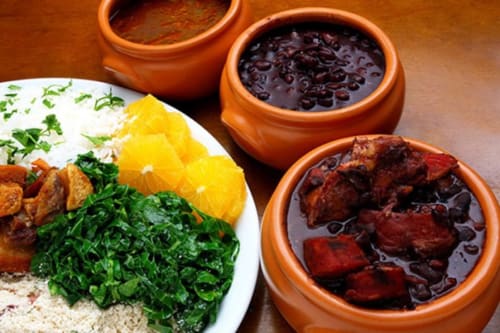 Feijoada brasileira tradicional com arroz, verduras, fatias de laranja e caldo de feijão preto em tigelas de terracota no Rio Vibes Hostel,