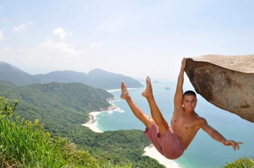 Jovem em equilíbrio sobre uma rocha de penhasco com vista para uma baía turquesa e montanhas florestadas, aventura no Rio de Janeiro