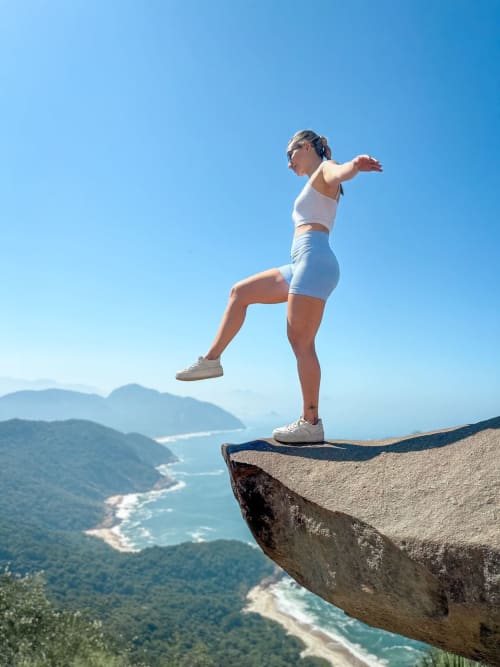 Mulher praticando yoga no topo de um penhasco com vista para as montanhas costeiras e o oceano do Rio de Janeiro