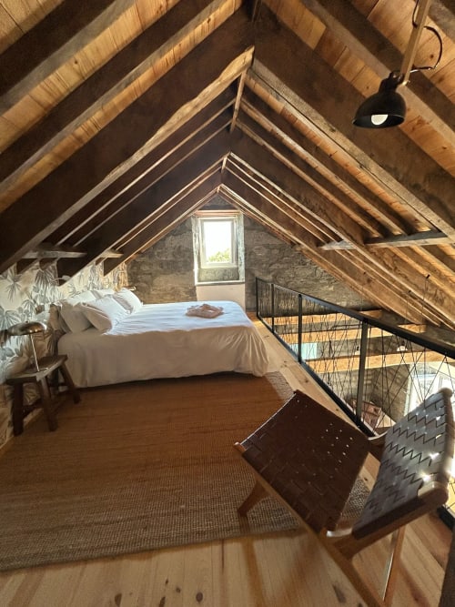 Quarto rústico no sótão da Cá Por Casa Pico com vigas de madeira à vista, cama branca, janela com vista para o mar e comodidades modernas