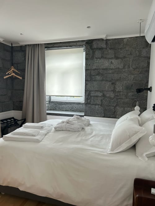 Quarto moderno com roupa de cama branca, paredes de pedra escura, janela com vista para o mar e decoração minimalista em Cá Por Casa Pico, Açores