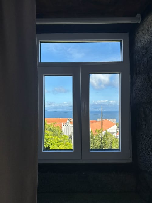 Janela moderna com vista para o mar sobre a ilha do Pico, telhados de cerâmica laranja e vegetação verde na Cá Por Casa Pico Açores