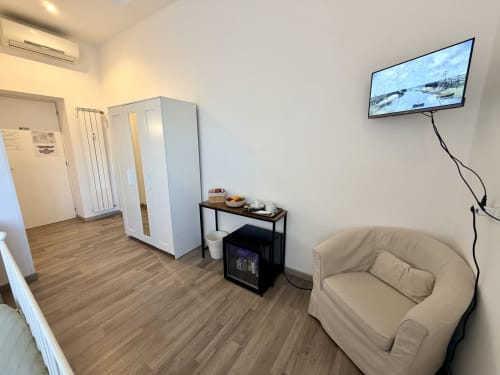 Accogliente camera moderna presso l'Enjoy Vatican B&B di Roma con poltrona beige, TV a schermo piatto, armadio e pavimentazione in legno