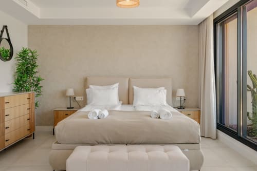 Nowoczesna sypialnia w apartamencie HomyMarbella w Mijas z beżowym łóżkiem, białymi poduszkami, drewnianymi stolikami nocnymi i widokiem na ogród