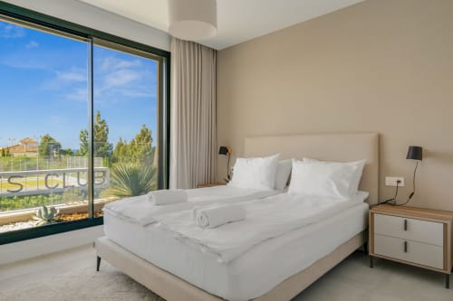 Nowoczesna sypialnia z widokiem na ogród w apartamencie HomyMarbella w Miјasie, z łóżkiem w białej pościeli i stolikami nocnymi