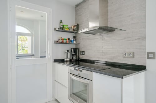 Nowoczesna biała kuchnia z czarnym blatem, urządzeniami ze stali nierdzewnej i szarą mozaiką w apartamencie HomyMarbella