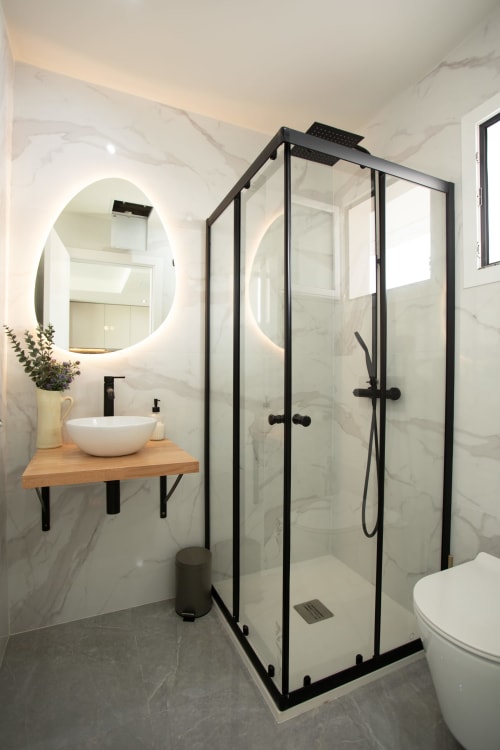 Baño moderno en Villa Gozamena con ducha de marco negro, paredes de mármol, espejo redondo iluminado, tocador de madera y