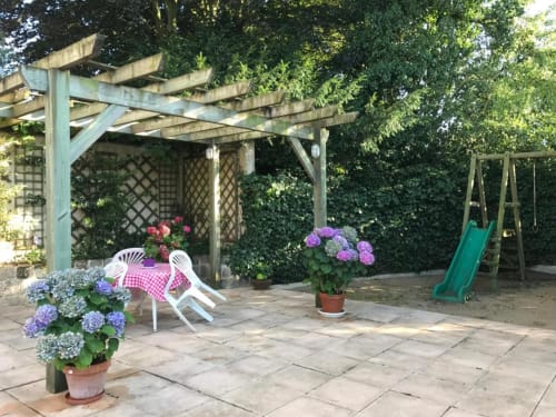 Terrasse du jardin La Pierre Levée avec pergola, hortensias fleuris, chaises blanches et balançoire pour enfants à La Gaubretière