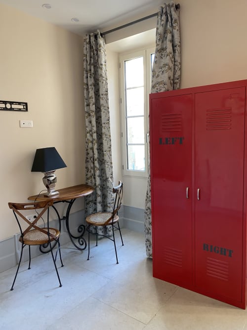 Le Clos Saint Maurin bedroom with red locker-style wardrobe, floral curtains, desk with lamp, and garden view door.