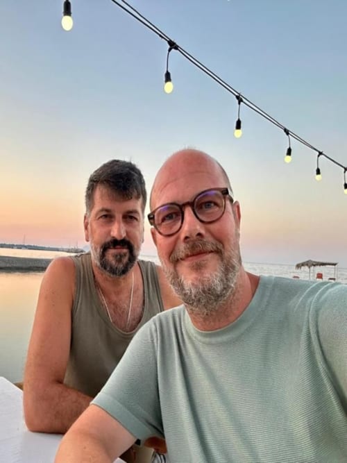 Two guests smile at Pink-Shell guesthouse in Santiago de Riba-Uí, Portugal, with evening string lights and waterfront view