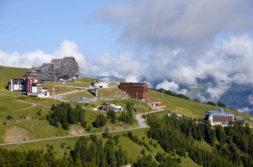 Station alpine L'Alp'In nichée dans les prairies verdoyantes, bâtiments montagnards et forêts de conifères à La Plagne Tarentaise