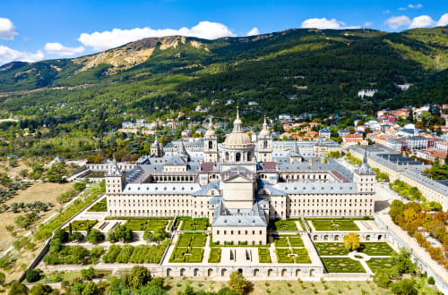 Vista aérea de un majestuoso monasterio con cúpula, jardines formales, montañas y el pueblo de Collado Villalba en el valle