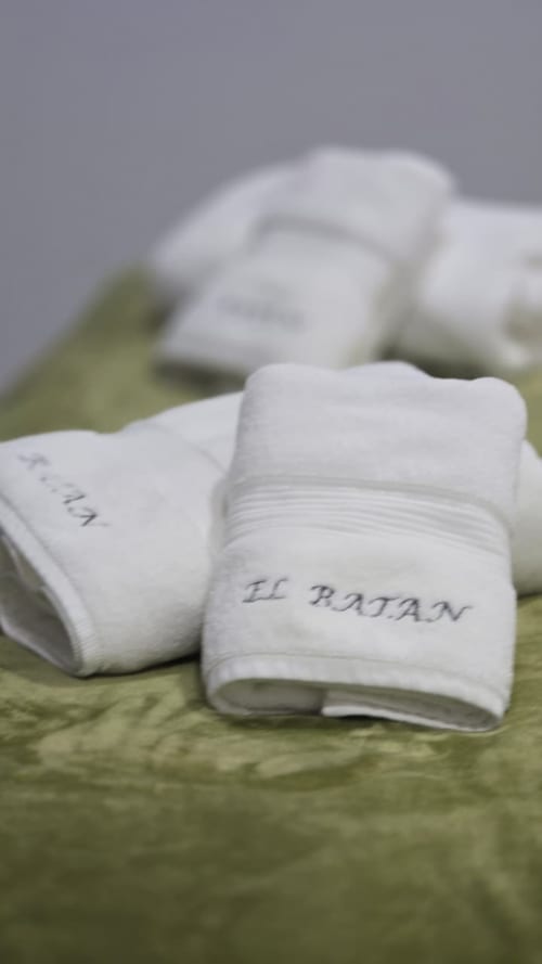 Zapatillas de spa blancas y mullidas con la marca del Hotel El Batán sobre toalla verde en hotel de lujo