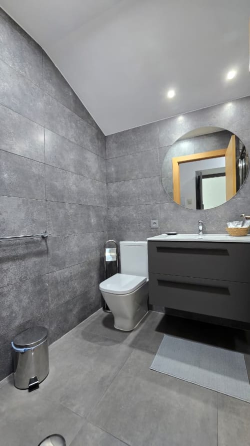 Baño moderno del Hotel Spa El Batán con paredes de azulejos grises, espejo redondo, vanidad negra e inodoro blanco en Castillo de