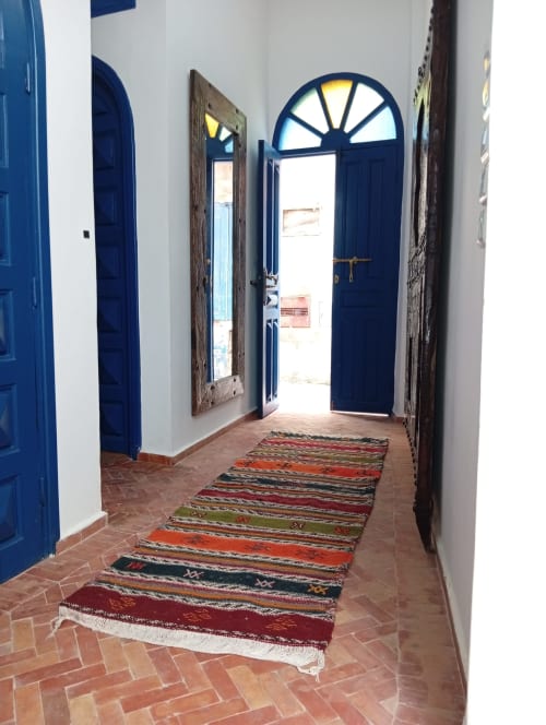 Couloir du Riad Le Village Mogador à Essaouira avec portes bleues, tapis rayé coloré et fenêtres cintrées