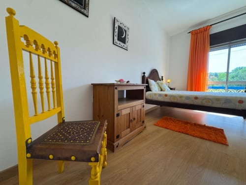 Quarto luminoso na Casa Amarela em Lagos com cadeira amarela, móveis em madeira, tapete laranja e vista da cidade pela janela