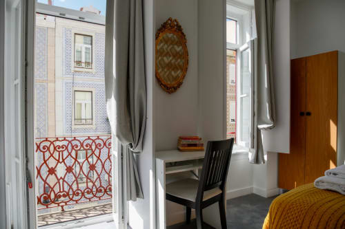 Bright Lisbon hostel room with desk, chair, balcony overlooking Portuguese architecture, wooden wardrobe and ornate mirror.