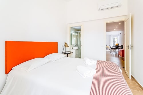 Quarto moderno com cabeceira laranja vibrante, roupa de cama branca e banco listrado no apartamento Barracuda em Algarve