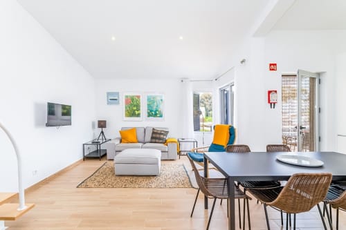 Moderno apartamento de praia Barracuda no Algarve com sala aberta, zona de refeições, sofá cinzento, cadeiras de madeira, TV e vista para a piscina