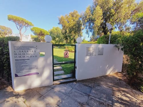 Entrada da piscina Algarve Beach Holidays com portão branco e sinalização, rodeada por árvores mediterrânicas