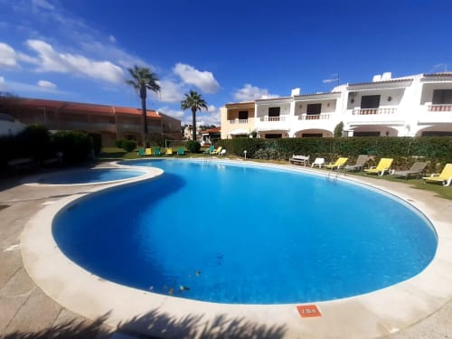 Grande piscina oval em Algarve Beach Holidays com espreguiçadeiras, palmeiras e edifícios brancos sob céu azul