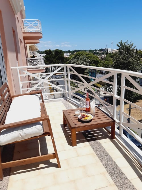 Terraço ensolarado na Algarve Beach Holidays com espreguiçadeira em madeira, mesinha de café, grades brancas e vista para jardins exuberantes