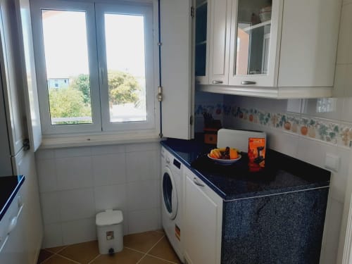 Cozinha moderna de apartamento com bancada escura, máquina de lavar, armários brancos e vista para a praia do Algarve