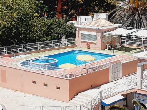 Área de piscina do Algarve Beach Holidays com piscina azul, guarda-sóis e edifício em terracota com vista para jardins tropicais
