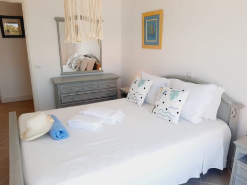 Quarto luminoso em Algarve Beach Holidays com cama branca, almofadas decorativas, cômoda cinza com espelho e tema costeiro