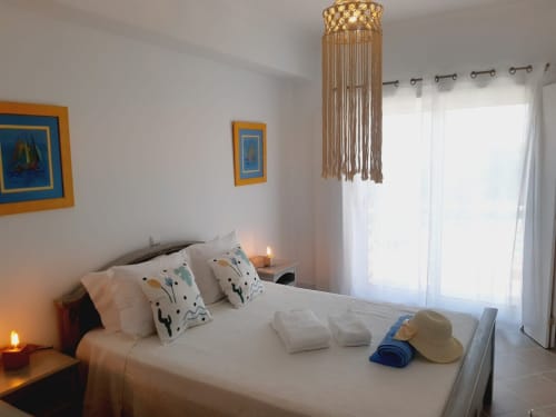Quarto luminoso no Algarve Beach Holidays com cama branca, almofadas decorativas, lustre de macramé e portas para pátio