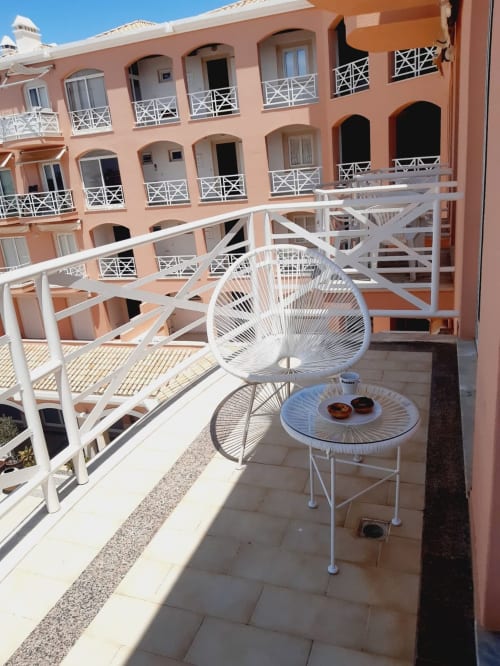 Terraço ensolarado do apartamento de praia VILLALUSA com cadeira branca, mesa e edifício do hotel em tons de coral ao fundo, Algarve,