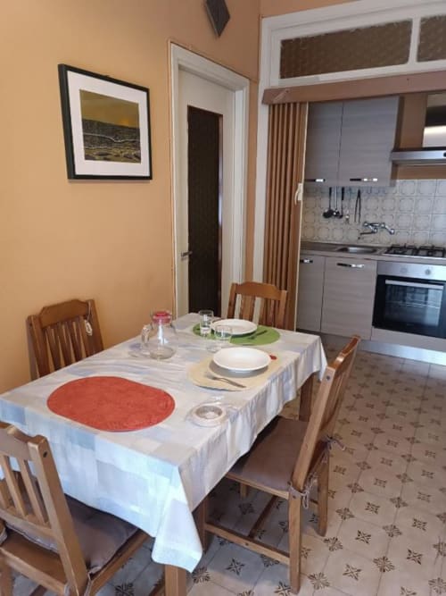 Zona pranzo dell'appartamento Residenza Adalgisa con tavolo apparecchiato, sedie in legno, cucina e quadri incorniciati a Finale Ligure.