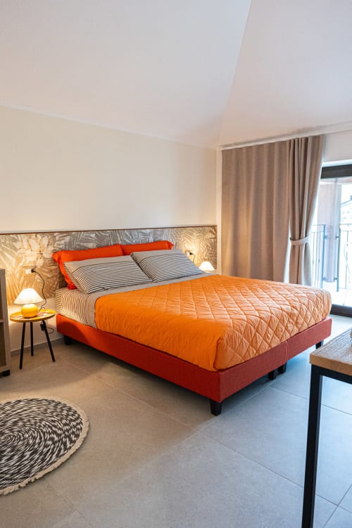 Camera Calendula presso AgriBio Villa Ribes con letto arancione-rosso, cuscini rigati, balcone con vista giardino, lampada moderna e