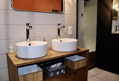 Modern bathroom vanity with two white round vessel sinks on wooden countertop, white tile backsplash, and orange-framed