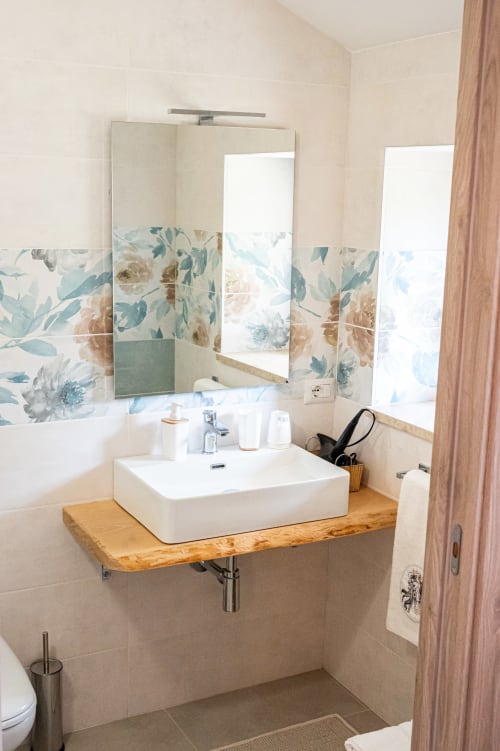 Bagno moderno nella suite Clematide di AgriBio Villa Ribes con lavabo bianco, ripiano in legno e rivestimento floreale