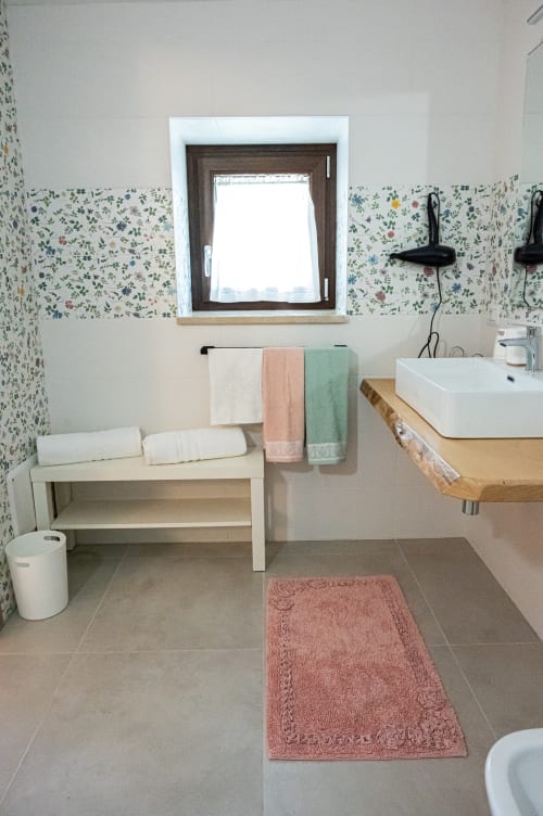 Bagno moderno di AgriBio Villa Ribes con carta da parati floreale, lavandino bianco, mobile in legno, finestra e tappetino da bagno rosa