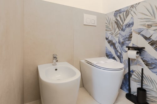 Moderno bagno nel loft Solumnia Milano con bidet e wc bianchi, accessori neri e decorazioni botaniche blu alle pareti
