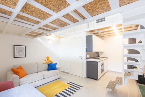 Moderna zona living in loft a Milano con divano beige, cucina a vista, travi in sughero e dettagli colorati presso Solumnia