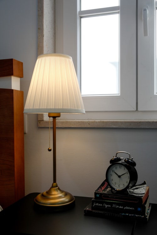 Brass table lamp with cream pleated shade on bedside table at Lisbon Poets Inn hostel, with alarm clock and books